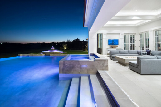 A shot of a patio with a firepit and TV on the right With a marble staircase on the left descending into a well-lit infinity pool.